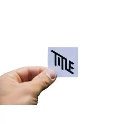 Hand holding a 1.5 inch TITLE MTB logo sticker in black on white background, part of a 6 pack set with 3 black and 3 white die-cut stickers.
