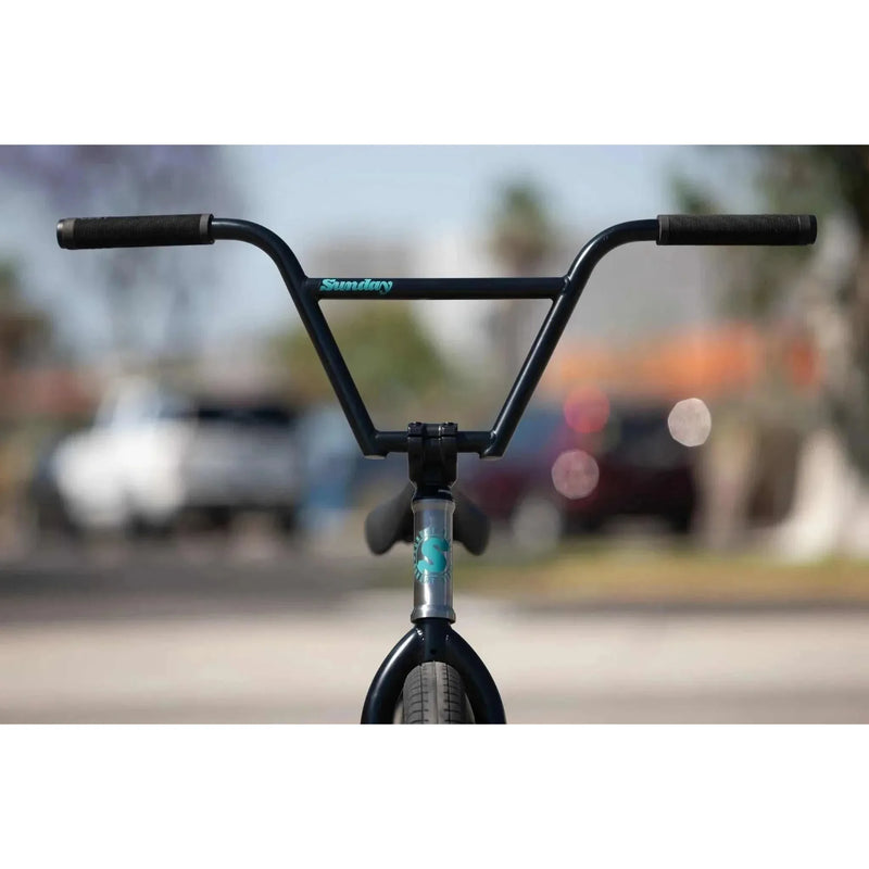 Front view of Sunday Forecaster 20" complete bike handlebar and fork with black grips and blue logo on a blurred outdoor background