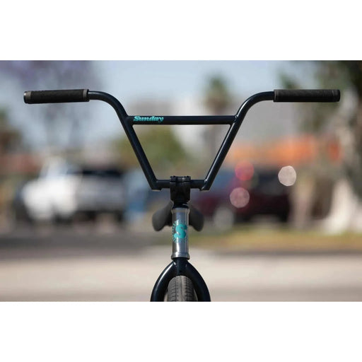Front view of Sunday Forecaster 20" complete bike handlebar and fork with black grips and blue logo on a blurred outdoor background