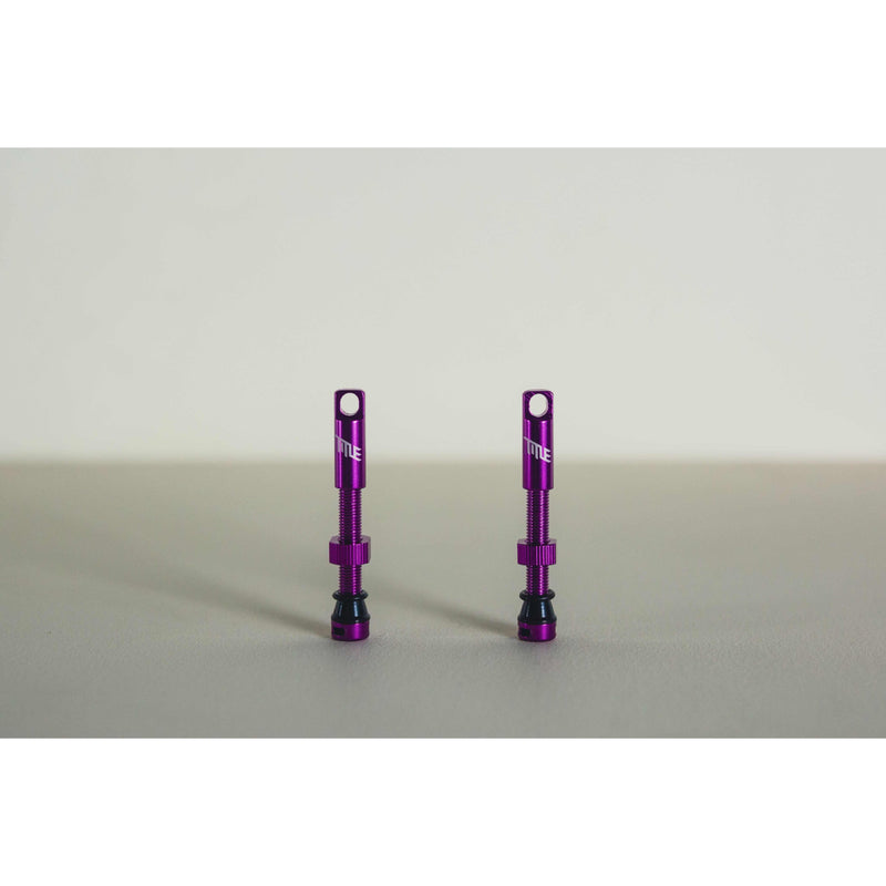 Pair of purple MTB tubeless presta valves with double air duct and metal caps on a beige background