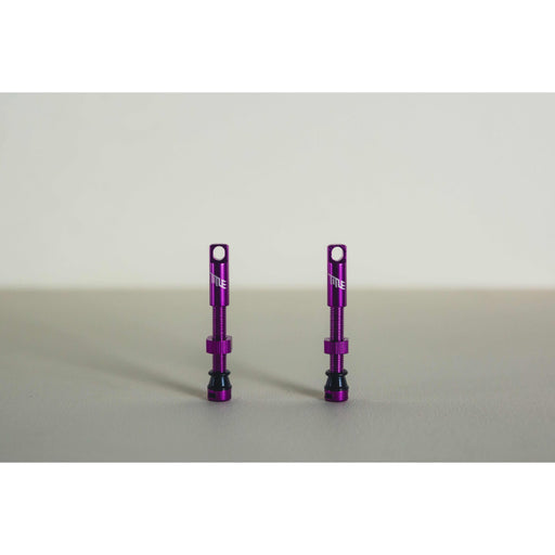Pair of purple MTB tubeless presta valves with double air duct and metal caps on a beige background